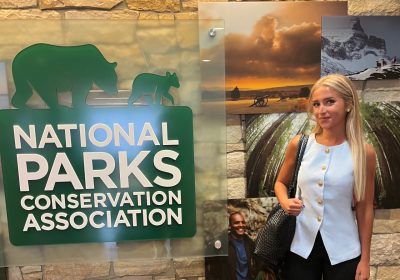 woman in light blue blouse with blonde hair stands next to a national parks conervation association sign