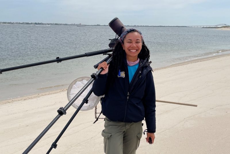 Woman carrying a spotting scope on a tripod over her shoulder while walking along a beach