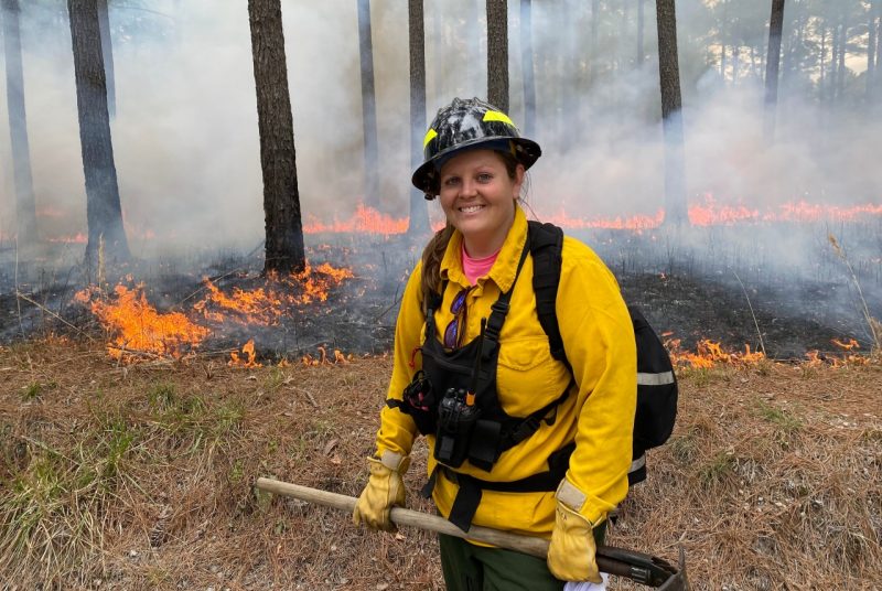 A woman wears protective clothing and a helmet and stands in front of a fire line amid pine trees.