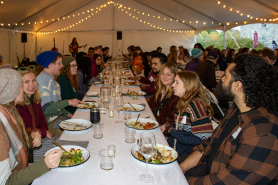 The photos shows three long dinner tables covered in white table cloths populated by students and faculty and alumni eating dinner and conversing.