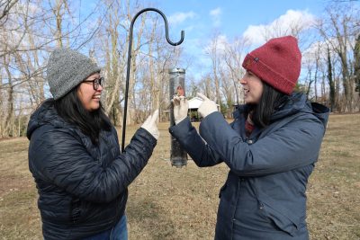 Sara Teemer and Dana Hawley, who is holding a house finch, are at a bird feeder that house finches frequent.